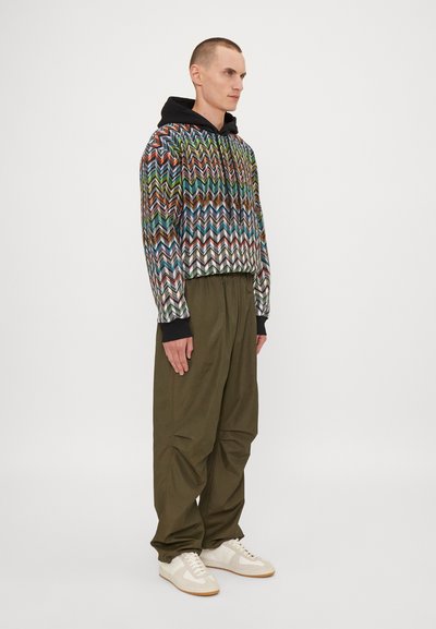 Bunt gewebte, Chevron-musterte, kurz geschnittene Kapuzenjacke mit schwarzer Kapuze, kombiniert mit weiten olivgrünen Hosen und weißen Sneakers. Glatte Textur.