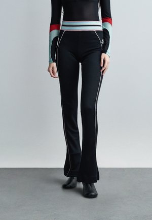 Osoba nosiaca čierne zvonové nohavice s bielymi bočnými švami a farebným pruhovaným pásom, v kombinácii s čiernymi topánkami a dlhým rukávom.