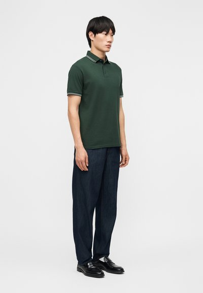 Homme debout portant un polo vert foncé avec bordure blanche, un pantalon ample foncé et des chaussures en cuir noir, sur un fond clair uni.