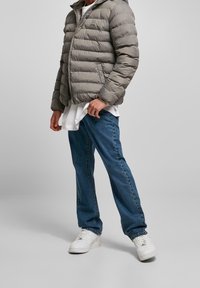 Person som bär en grå pufferjacka, vit skjorta, blå jeans och vita sneakers, stående mot en enkel grå bakgrund.