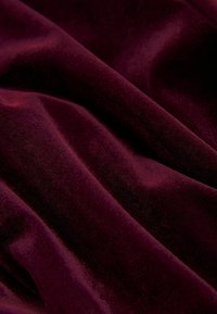 Tissu en velours bourgogne avec une texture douce, affichant des plis lisses et fluides ainsi qu'une couleur riche et profonde. La lumière se reflète subtilement sur la surface.