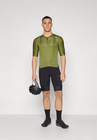 Grünes kurzärmeliges Fahrradjersey mit einem Frontreißverschluss, kombiniert mit schwarzen Shorts. Das Modell hält einen schwarzen Helm und trägt Fahrradschuhe und -socken.
