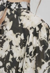 Blusa a motivi in bianco e nero con tessuto semi-trasparente, maniche plissettate e un colletto alto con due bottoni neri sul retro.