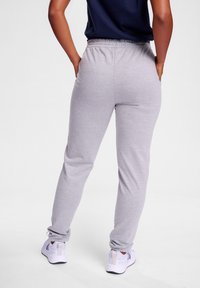 Osoba stojąca, ubrana w jasno szare spodnie dresowe, białe trampki i ciemnoniebieską górę, ręce spoczywają na biodrach, odwrócona tyłem.