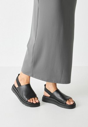 Sandali neri in pelle con design a punta aperta, cinturino alla caviglia e suola in gomma testurizzata; indossati con un lungo vestito grigio.
