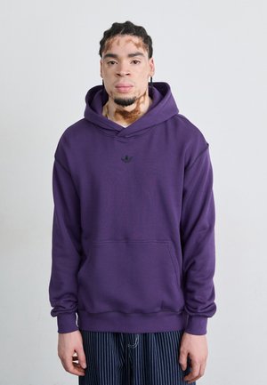 Sweat à capuche violet avec une poche kangourou et des coutures noires. Petit logo noir sur la poitrine. Le modèle porte un pantalon rayé.