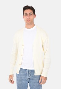 Cardigan giallo chiaro realizzato in tessuto a maglia, con scollatura a V, cinque bottoni e polsini a costine. Indossato sopra una maglietta bianca e jeans.
