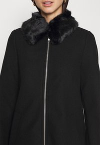 Cappotto nero con colletto in pelliccia sintetica, chiusura con zip e maniche lunghe. Tessuto strutturato con finitura liscia. Nessun motivo o decorazione visibile.