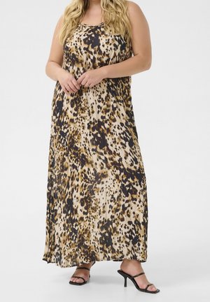 Vrouw met een mouwloze maxijurk met luipaardprint en zwarte hoge sandalen met bandjes, staand met gevouwen handen.
