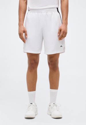 Férfi fehér atlétikai rövidnadrágban fekete csíkokkal, fehér zokniban és fehér sportcipőben, egyszínű világos háttér előtt áll.