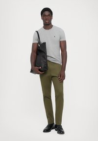 Grå kortærmet t-shirt, olivengrønne bukser og sorte lædersko. Sort skuldertaske med minimalistisk design, holdt med højre hånd.