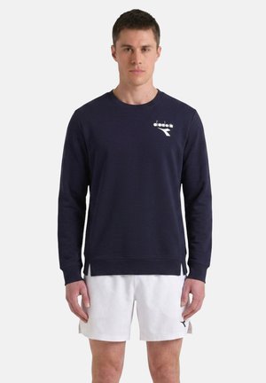Giovane uomo con felpa blu navy a maniche lunghe e pantaloncini sportivi bianchi in piedi, rivolto frontalmente su uno sfondo uniforme.