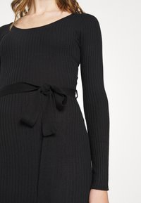 Robe noire en tricot côtelé avec manches longues, encolure dégagée et ceinture en tissu à nouer à la taille. Texture lisse et coupe ajustée.