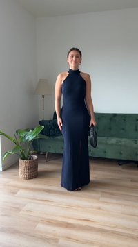 Morska halter obleka s prilegajočo silhueto in stranskim razporkom. Obuta z črno torbico, na ozadju zelene sedežne garniture in svetlega lesenega poda.