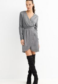 Vestido cruzado gris metálico con mangas largas, cintura entallada y dobladillo corto y ligeramente asimétrico. Combinado con botas negras hasta el muslo.