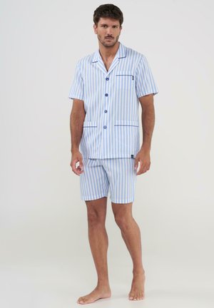 Hombre de pie descalzo vistiendo pijama de camisa de manga corta con botones y pantalones cortos a rayas verticales en azul claro y blanco.