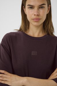 Ung kvinne med rett brunt hår som har på seg en mørk lilla skjorte med en liten brodert logo, armene krysset over brystet.