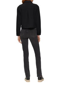 Vrouw in een zwarte cropped jas, donkergrijze slim-fit jeans en zwarte sneakers met beige zolen, gezien vanaf de achterkant op een witte achtergrond.