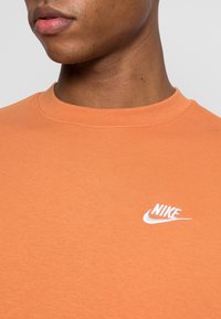 Camiseta de algodón naranja con cuello redondo acanalado, que presenta un logo de Nike bordado en blanco en el lado izquierdo. Textura suave.
