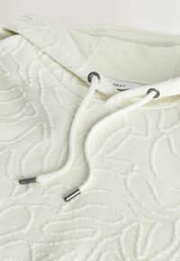 Sweat à capuche blanc texturé avec motif floral en relief, cordons blancs avec embouts en métal, et une étiquette de marque "NEXT" visible à l'intérieur du col.