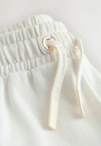 Nahaufnahme eines weißen elastischen Bunds mit Kordelzug, der durch eine goldene Metallöse auf weichem Stoff gezogen ist.