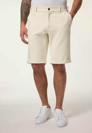 Mand iført opkrymmede beige shorts, hvide sneakers, med tatoveret venstre arm og hænderne i lommerne, stående på en neutral baggrund.