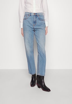 Femme portant un jean taille haute bleu clair avec jambes droites, une chemise blanche boutonnée et des bottines noires avec des semelles roses.
