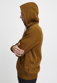 Hoodie marron avec une poche kangourou, capuche avec cordon de serrage ajustable et ourlet côtelé. Le tissu semble doux avec une texture lisse.