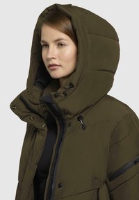 Giacca imbottita verde oliva con un grande cappuccio, zip frontale e bottoni a pressione. Presenta dettagli neri e un design slanciato.