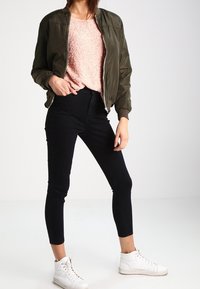 Femme portant une veste aviateur vert olive, un pull rose texturé, un jean noir moulant et des baskets montantes blanches, posant avec une main dans la poche.