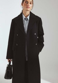 Melns dubultā krūšu virsjakas ar lieliem atlokiem, pogu detaļām un sānu kabatām. Valkāts virs gaiši zilas svītrainas krekla, turēts ar melnu somu.