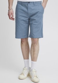 Light blue shorts, structured fabric, flat front with two side pockets, mid-length. Paired with white socks and beige sneakers.