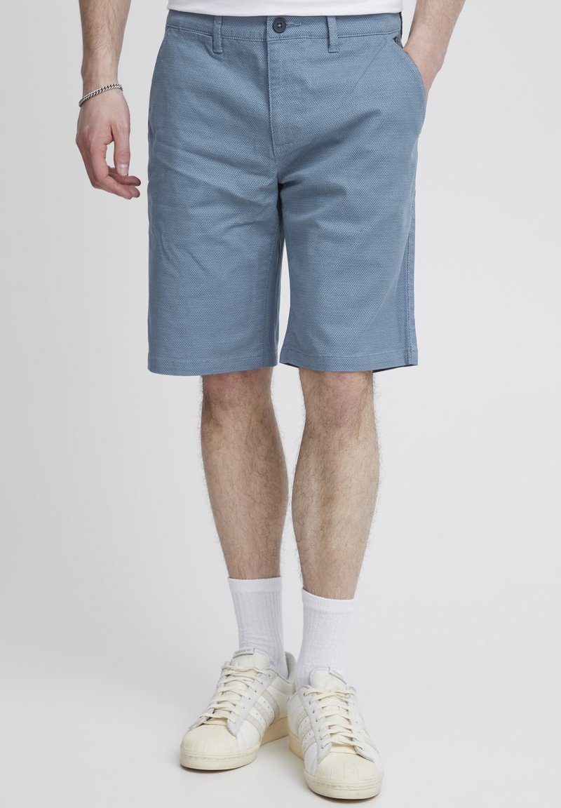 Light blue shorts, structured fabric, flat front with two side pockets, mid-length. Paired with white socks and beige sneakers.