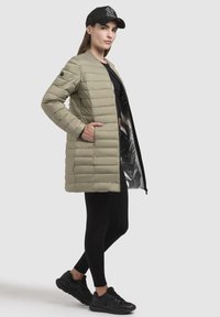 Puffjacka i olivgrönt, med ett kviltat mönster, långa ärmar, sidofickor och en reflekterande insida. Buren med svarta leggings och sneakers.