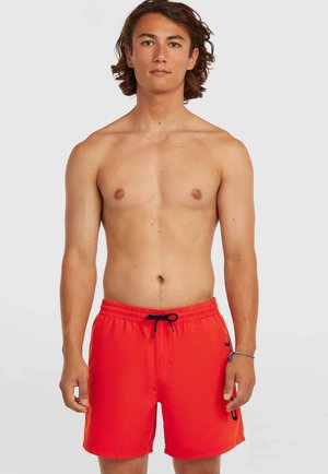 Man zonder shirt met halflang bruin haar, gekleed in felrode zwemshorts met een zwarte trekkoord, staand tegen een effen lichte achtergrond.