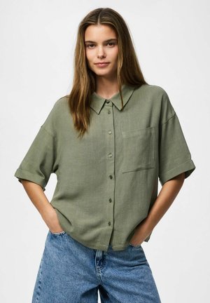 Jeune femme aux longs cheveux portant une chemise ample boutonnée vert olive et un jean bleu, les mains dans les poches, expression faciale neutre.