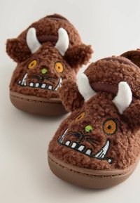 Chaussons pour enfants en peluche marron avec des visages de monstres brodés, des yeux orange, des nez verts, des cornes blanches et des dents proéminentes sur une surface claire.