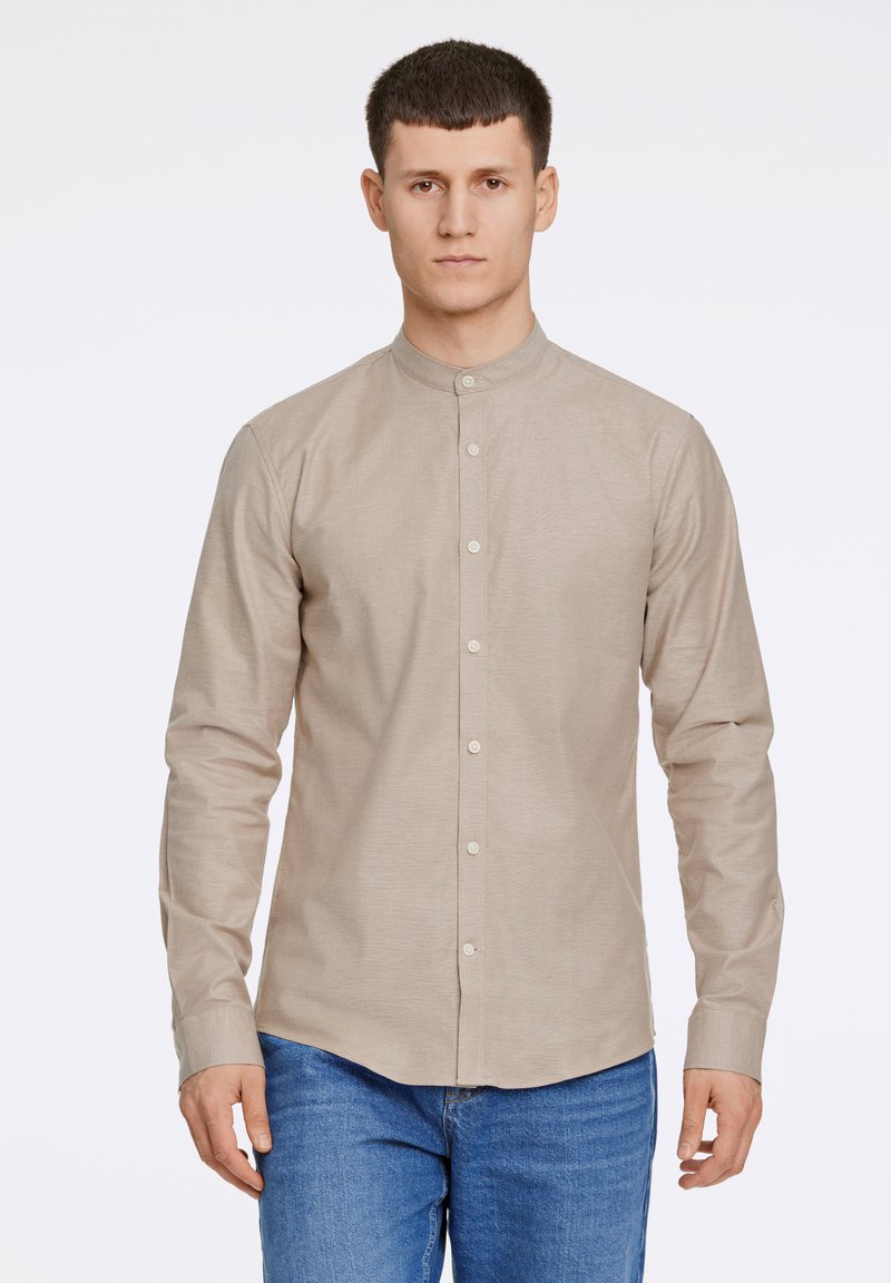 Μπεζ πουκάμισο με μακριά μανίκια και γιακά μανταρίνι, με διακριτική υφή και κούμπωμα με κουμπιά μπροστά· συνδυασμένο με μπλε τζιν.