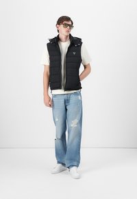 Vesta neagră căptușită cu glugă, fermoar frontal și logo; asortată cu un tricou gri deschis și blugi ușor distruși de culoare albastru deschis. Tenisi albi.