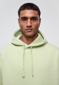 Lichtgroene hoodie van zacht materiaal, met een capuchon met trekkoord en een relaxte pasvorm. De textuur is glad zonder zichtbare patronen.