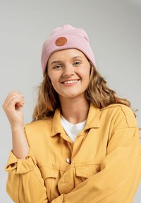 Różowa dziana czapka z teksturowanym wzorem i okrągłą skórzaną łatą; noszona z żółtą kurtką na białej koszuli.