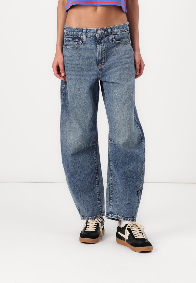 Personne portant un jean bleu ample, des baskets noires et blanches, et un haut court rayé bleu et rouge sur un fond uni.