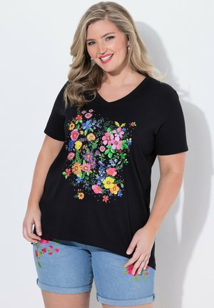 Kvinde smilende, iført en sort T-shirt med V-hals og farverigt blomsterprint samt lyseblå cowboyshorts med pink blomsterbroderi.