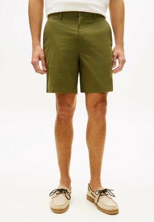 Mężczyzna w oliwkowo-zielonych spodenkach do kolan i beżowych mokasynach w stylu moccasin z ciemnymi sznurowadłami, stojący na jasnym tle.