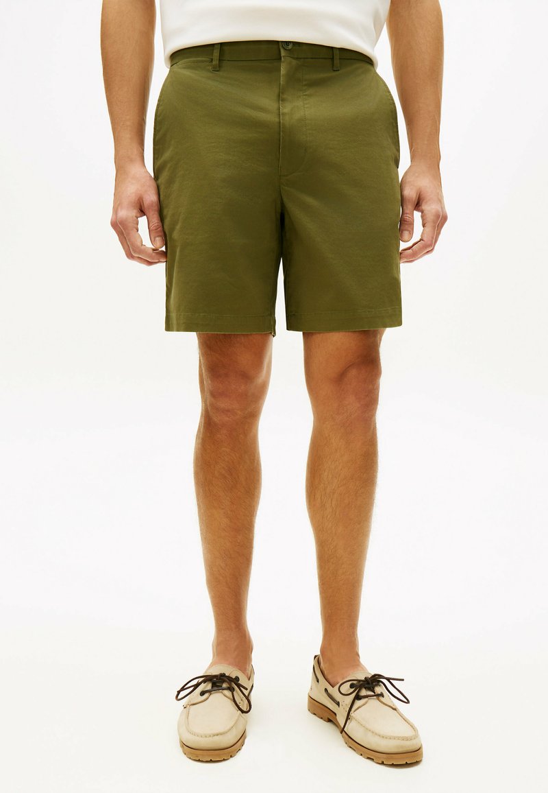 Man wearing olive green knee-length shorts and beige moccasin-style shoes with dark laces, standing against a light background.