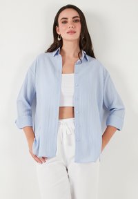 Light blue button-up shirt with a textured fabric, long sleeves rolled back, and a relaxed fit. Worn over a white top and light pants.
