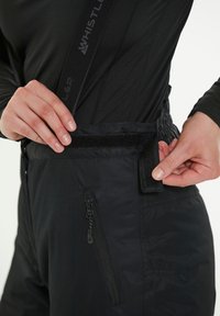 Mustat lasketteluhousut säädettävillä olkaimilla, joissa on tarranauha ja sivutasku vetoketjulla. Sujuva, vedenkestävä kangas.