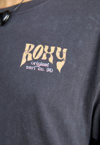 Mørk grå t-skjorte laget av myk bomull, med en falmet gul "ROXY"-logo og teksten "original surf co. 90" trykket på forsiden.