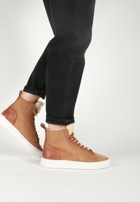 Ruskeat korkeat lenkkitossut, joissa on nahka- ja tekstiilipäälliset, valkoisella kumipohjalla ja karvoilla tehostettuna kauluksessa. Mustat käännetyt farkut.