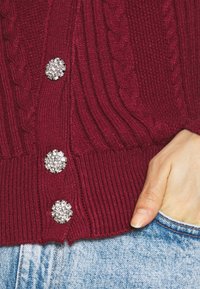 Burgundské pletené kardigán s ribbovanou strukturou, dekorativními kulatými kamínkovými knoflíky a lehce zkráceným střihem v pase.